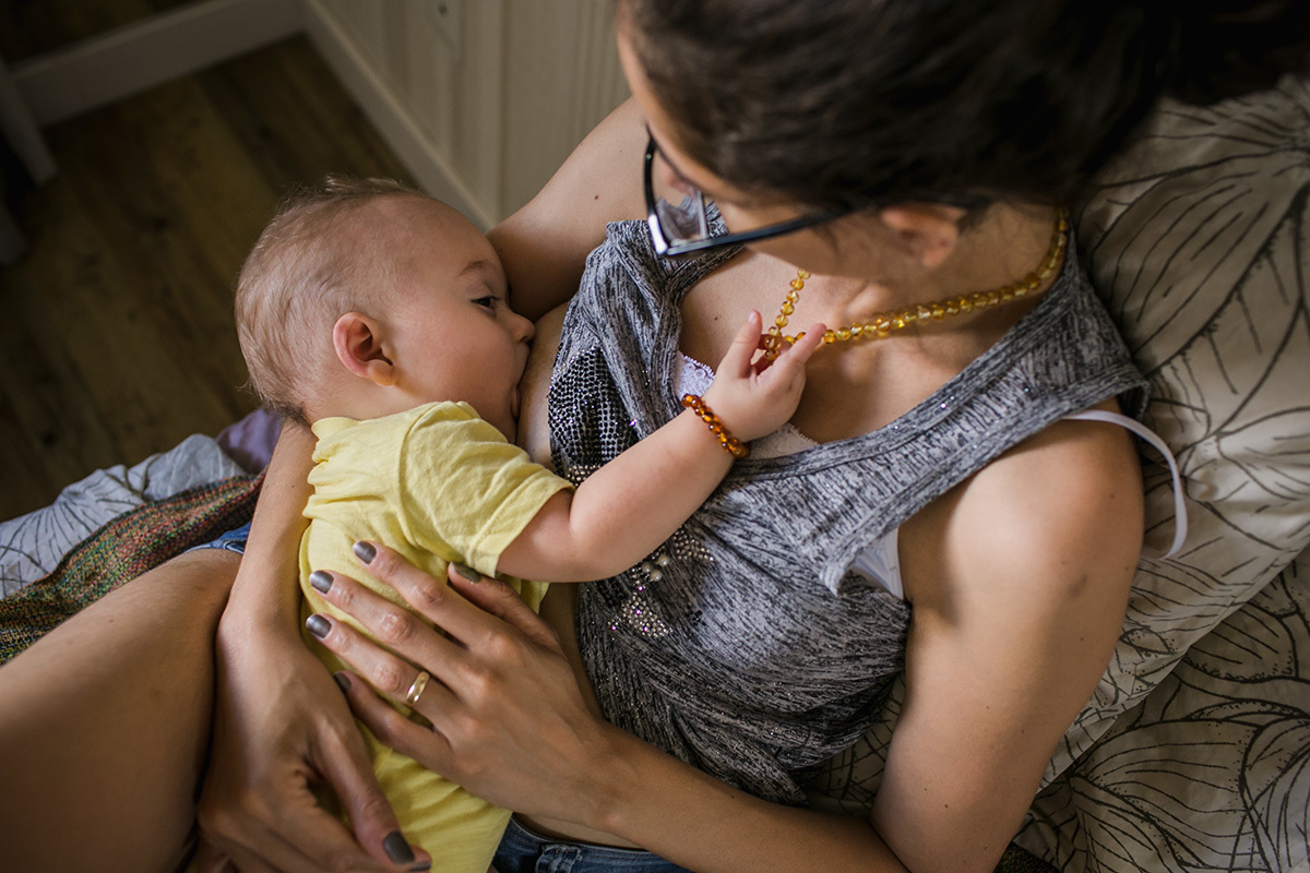 bebê  brincando com o colar de âmbar da mãe enquanto amamenta, por Adriana Costa