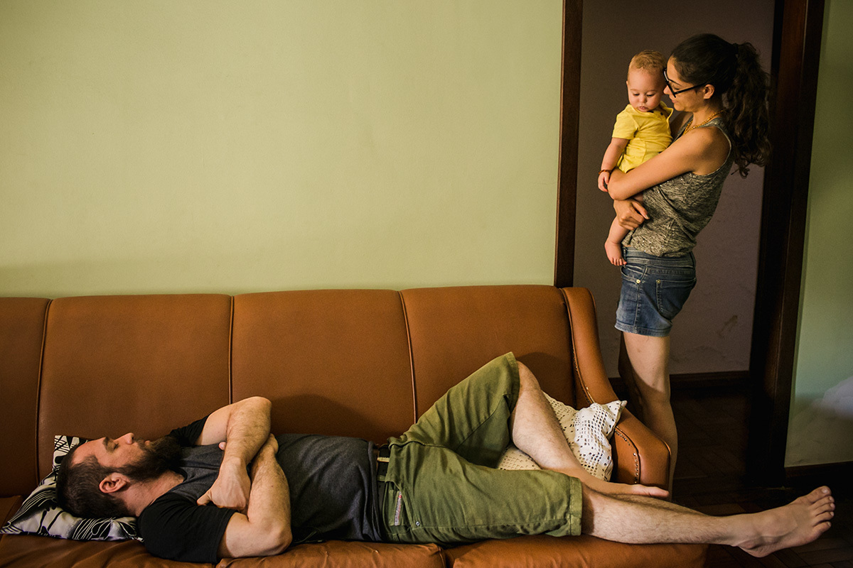 mãe e seu bebê no colo vendo o pai dormindo, em foto de Adriana Costa