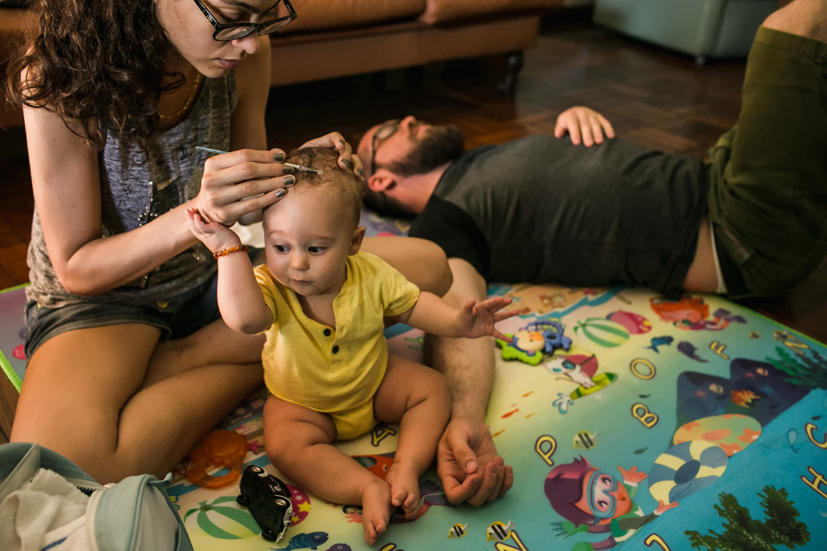 Foto de Adriana Costa da mãe penteando o cabelo do seu bebê enquanto ele empurra sua mão e o pai ao lado deles dormindo