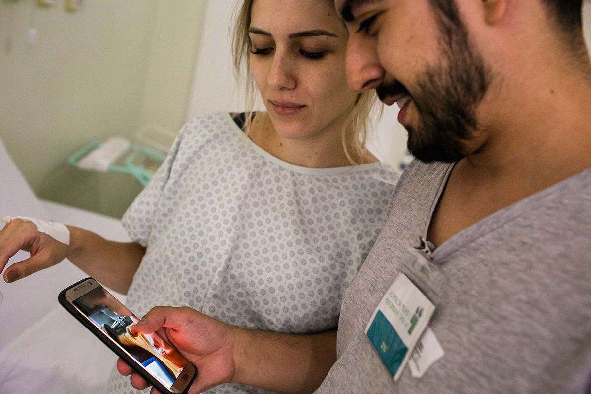 pais na maternidade para receber seu bebê e vendo o outro filho em casa pelo celular, por Adriana Costa