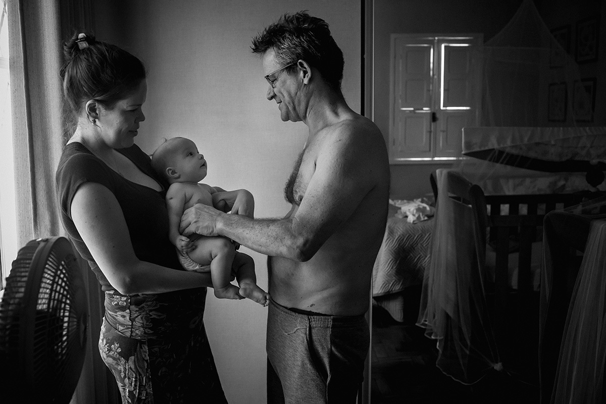 pai pegando sua bebê do colo da mãe para levar ao banho de sol, por Adriana Costa