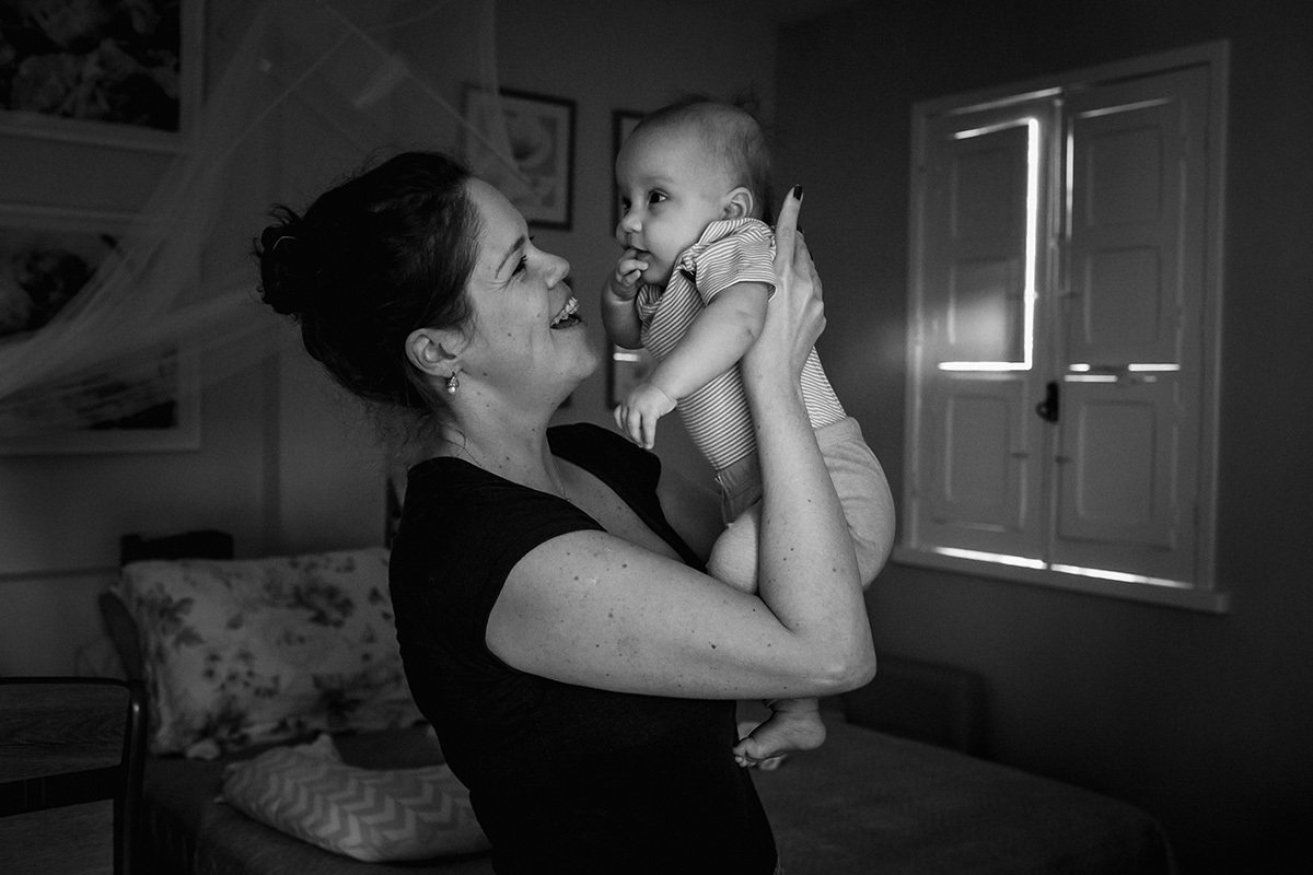 mãe admirando sua bebê de cinco meses sob uma bela luz de seu quarto, por adrianacostafotos