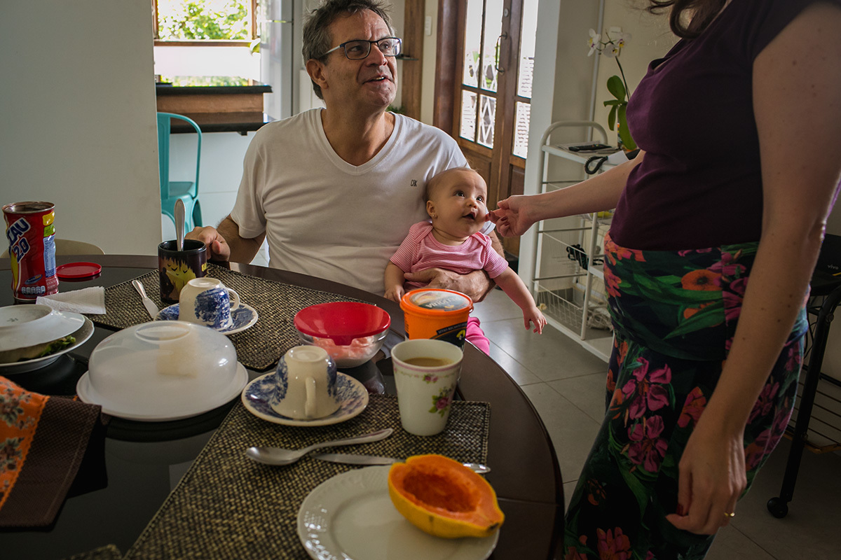mãe fazendo carinho na filha que está no colo do pai durante o café da manhã, por Adriana Costa