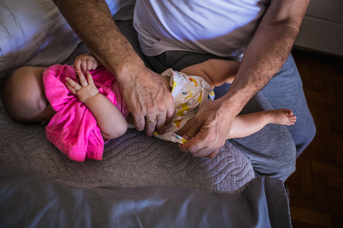pai trocando a fralda ecológica da filha enquanto ela esconde o rostinho sob a roupa, por Adriana Costa