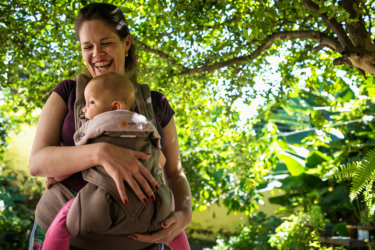 mãe feliz com sua bebê no canguru sob a sombra de árvores, por Adriana Costa