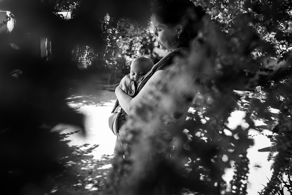 bebê no canguru para bebês junto a mãe adormece sobre a sombra de árvores, em foto de Adriana Costa