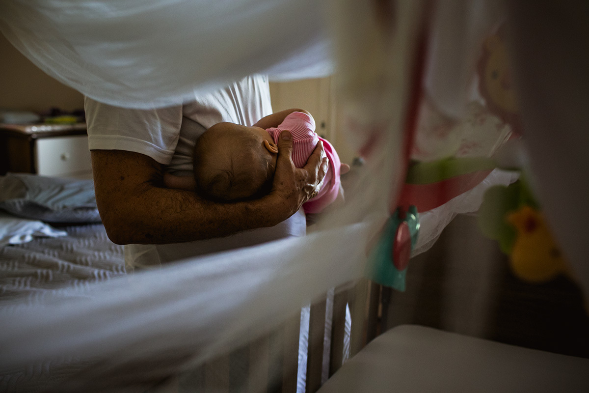 pai indo colocar a filha que dormiu no berço em um registro documental de Adriana Costa