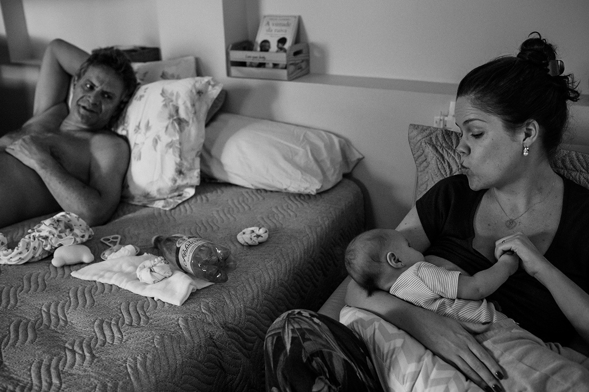 marido observa a esposa mandando beijo para a filha enquanto amamenta durante um acompanhamento documental da família feito por Adriana Costa