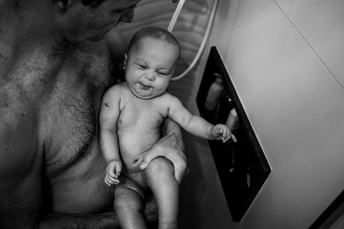 foto de Adriana Costa do bebê no colo do pai fazendo careta sob a água do chuveiro