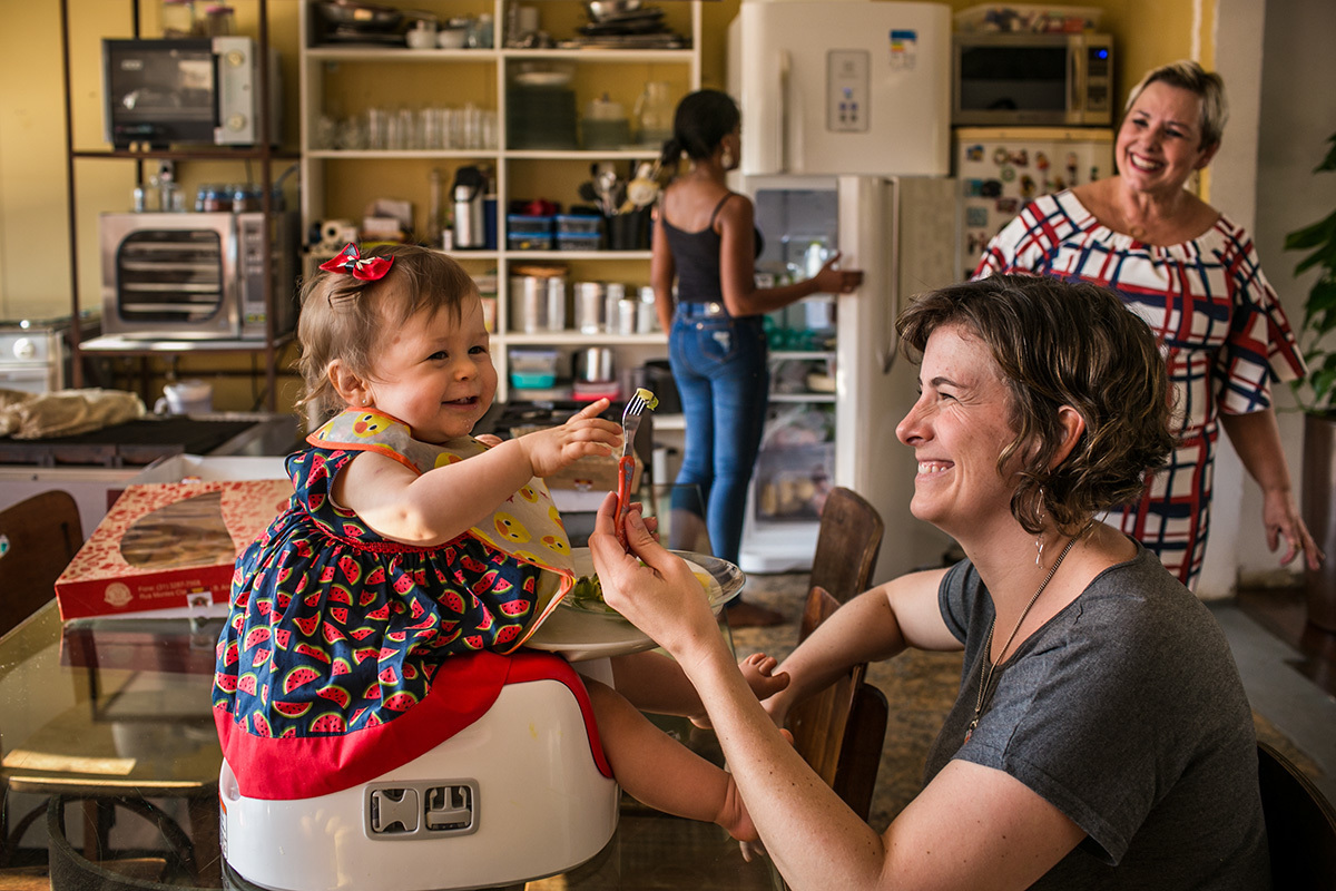 bebê rindo com sua mãe e avó enquanto come a fruta sobre a mesa da cozinha, por adrianacostafotos