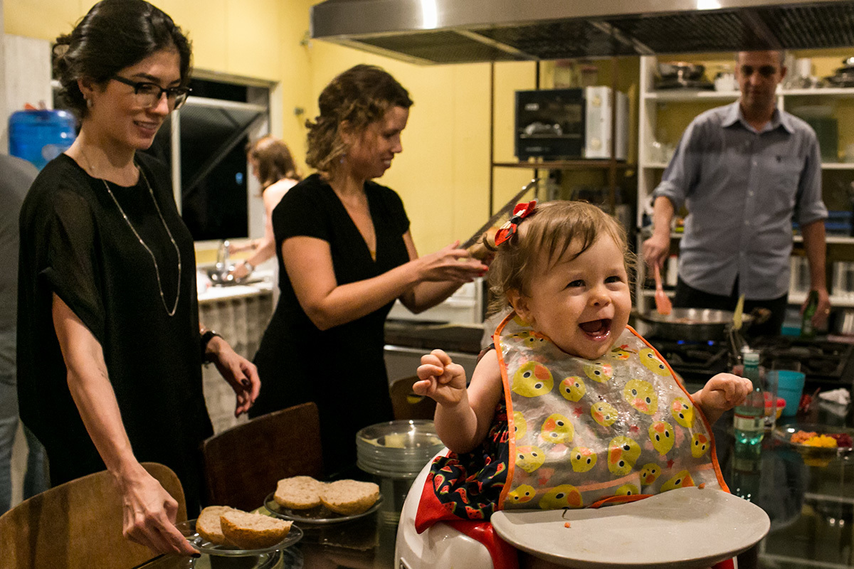 aniversariante de um ano dando risada enquanto janta na cozinha de sua avó rodeada por seus convidados, em foto de Adriana Costa