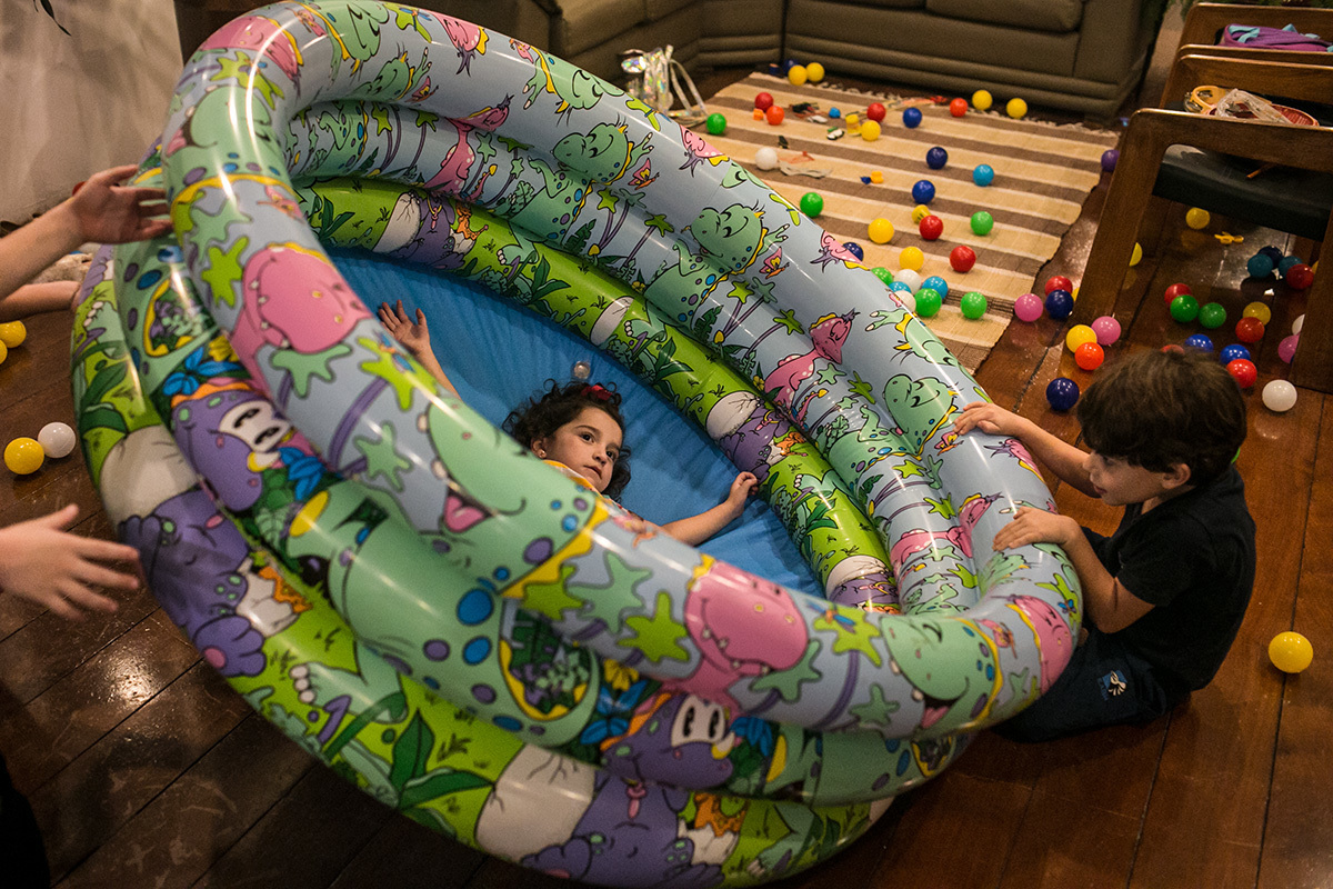 Meninos rodando uma piscina de bolinhas com outra criança dentro na maior diversão, por Adriana Costa