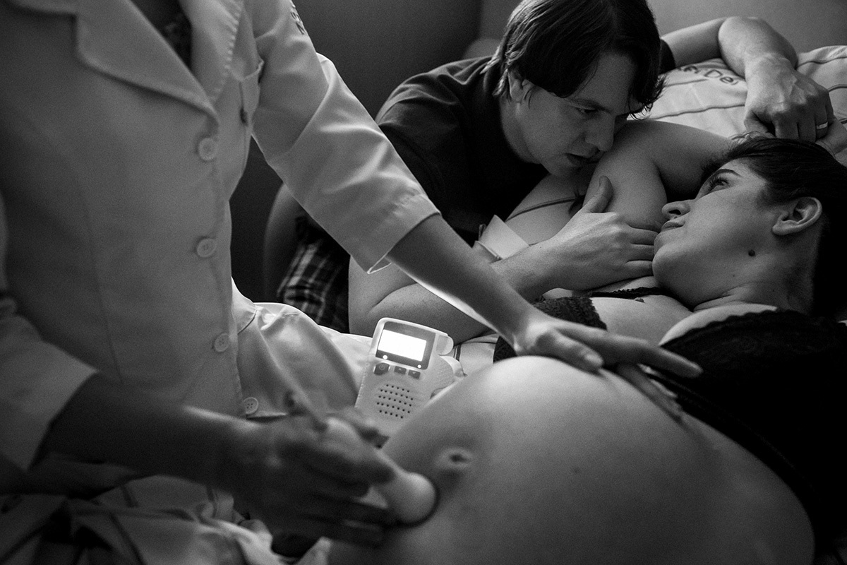 esposa e marido se entreolhando enquanto a enfermeira obstétrica avalia os batimentos cardíacos do bebê durante o trabalho de parto, em uma foto de Adriana Costa