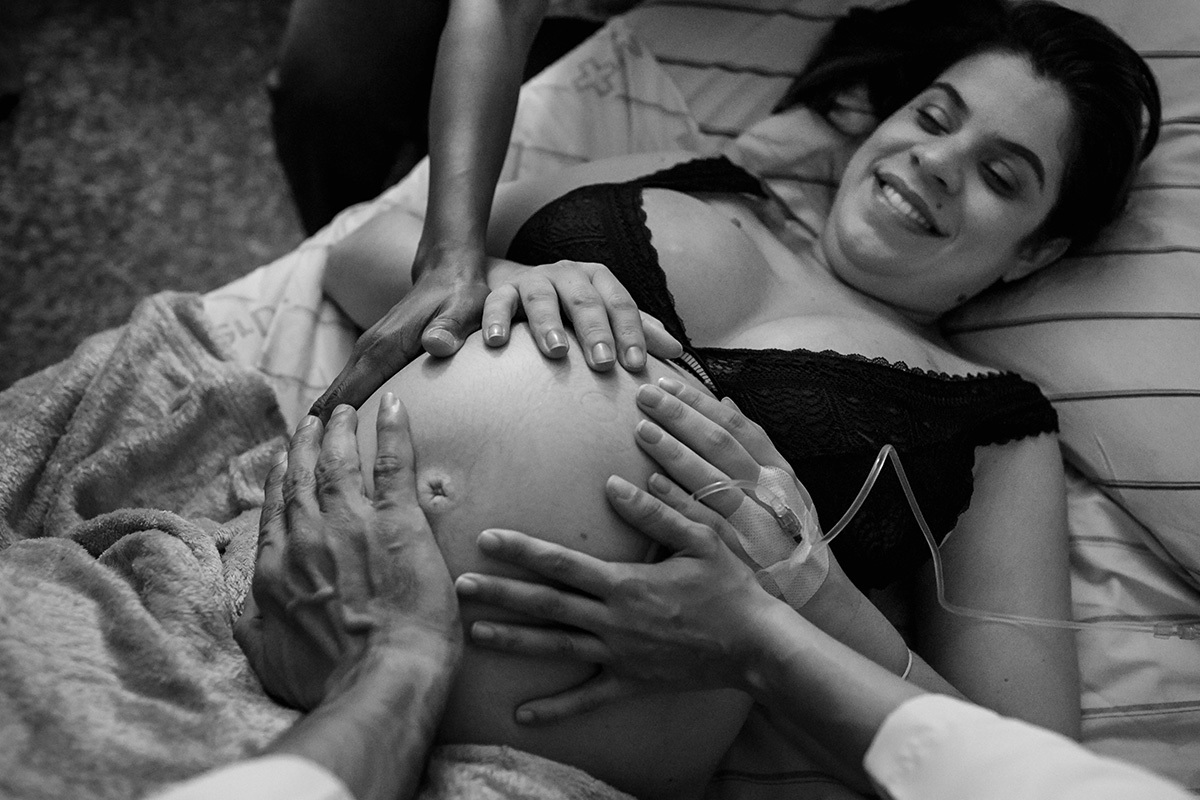 equipe médica com as mãos na barriga da gestante abençoando o bebê durante o trabalho de parto, por Adriana Costa