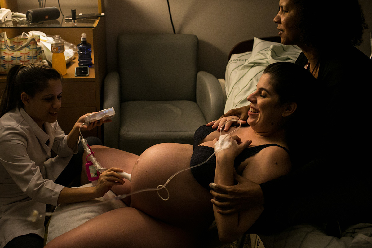 gestante apoiada em sua doula enquanto a enfermeira obstétrica avalia os batimentos cardíacos do bebê, por Adriana Costa
