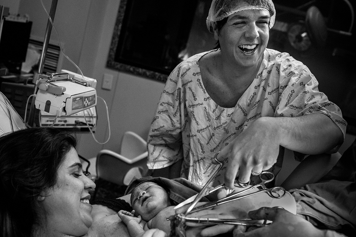 pai feliz e realizado ao cortar o cordão umbilical de seu primeiro filho, por Adriana Costa