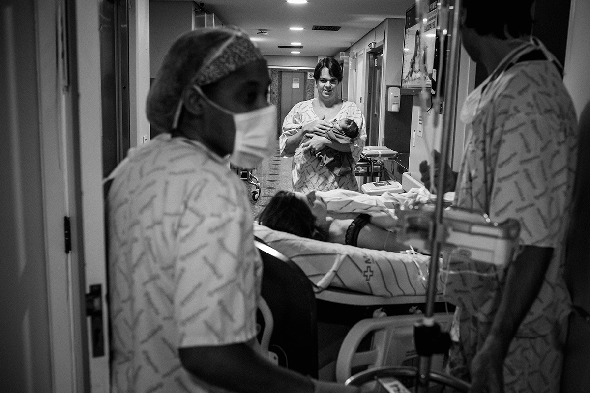 pai carrega seu filho enquanto sua esposa é transferida do bloco cirúrgico para o quarto, após o nascimento 