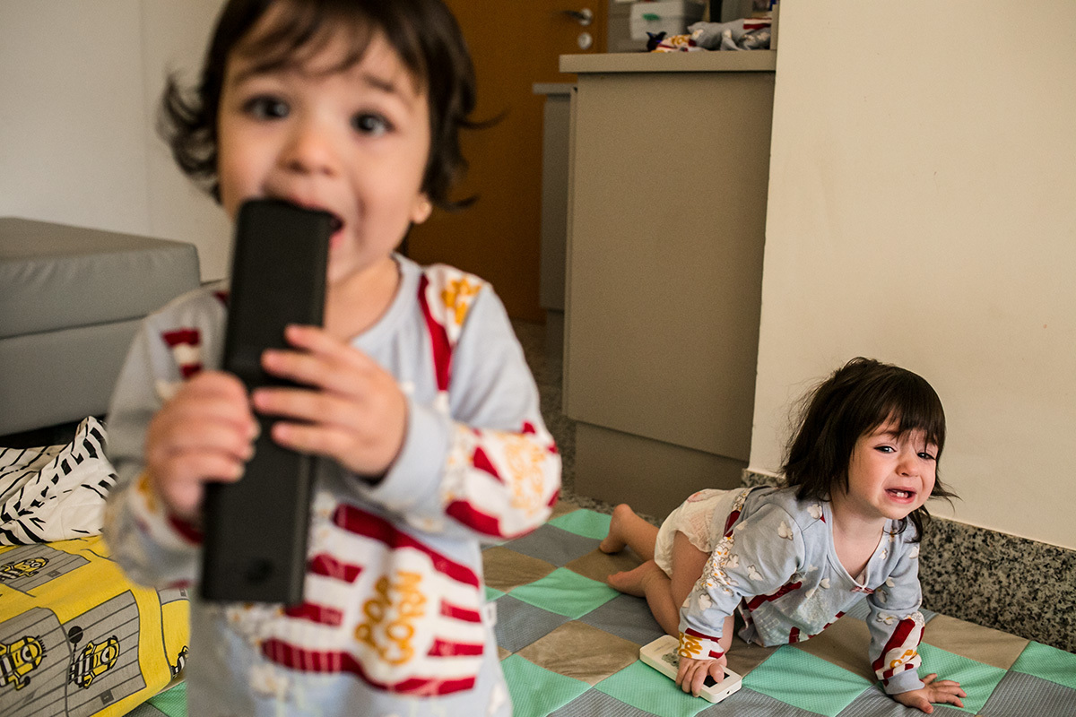 bebê mordendo controle remoto enquanto sua irmã gêmea engatinha chorando, em foto de Adriana Costa