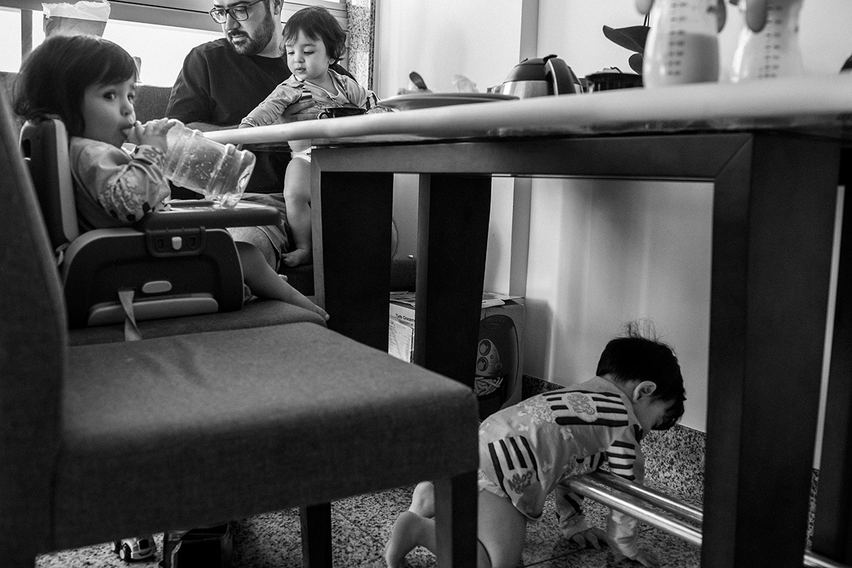 pai segura um de seus trigêmeos no colo durante o café da manhã enquanto a outra está na cadeirinha e o outro debaixo da mesa, por Adriana Costa