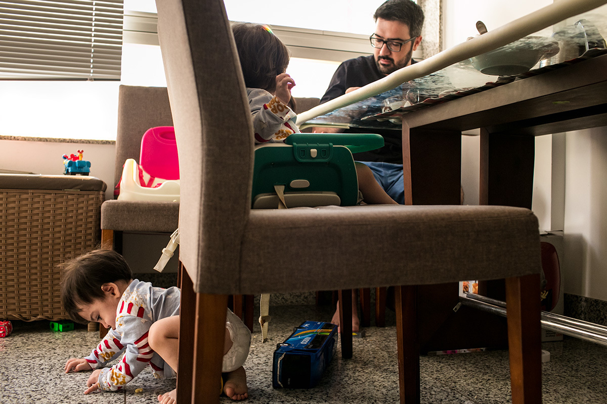 pai tomando café da manhã enquanto um de seus trigêmeos brinca debaixo da cadeira da mesa, em foto de Adriana Costa