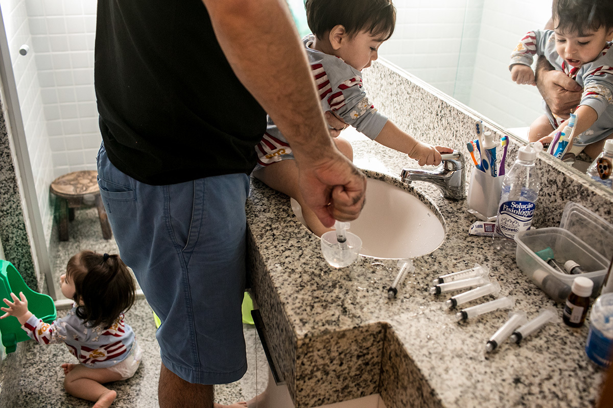 pai prepara a seringa com soro fisiológico para limpar o nariz de seus filhos gêmeos, em foto de Adriana Costa