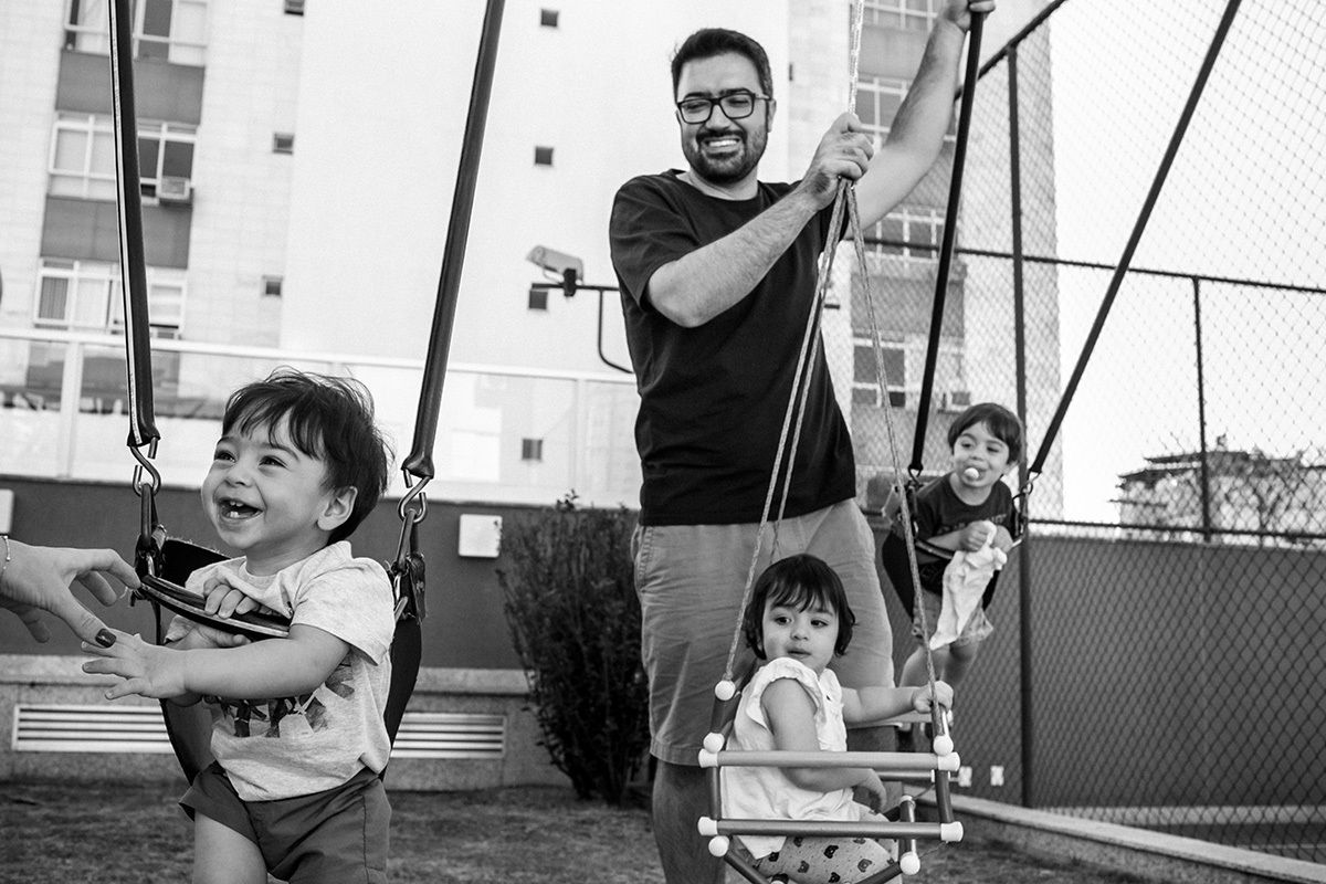 pai ri ao ver seus filhos se divertirem com sua esposa, Renata Lucena, no balanço do playground do prédio, em foto de Adriana Costa