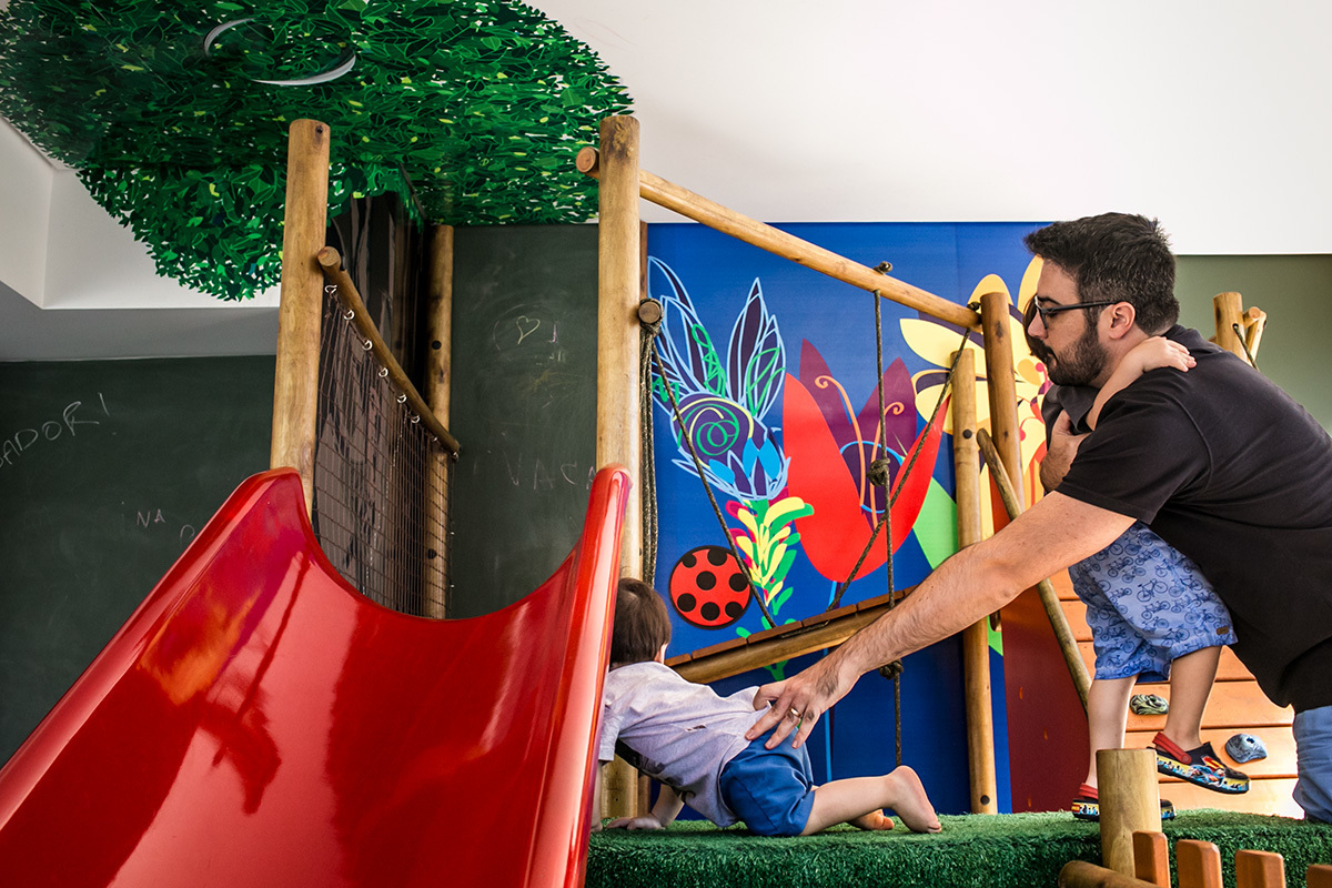 registro documental de Adriana Costa do pai tentando segurar seus dois filhos em um brinquedo de seu prédio