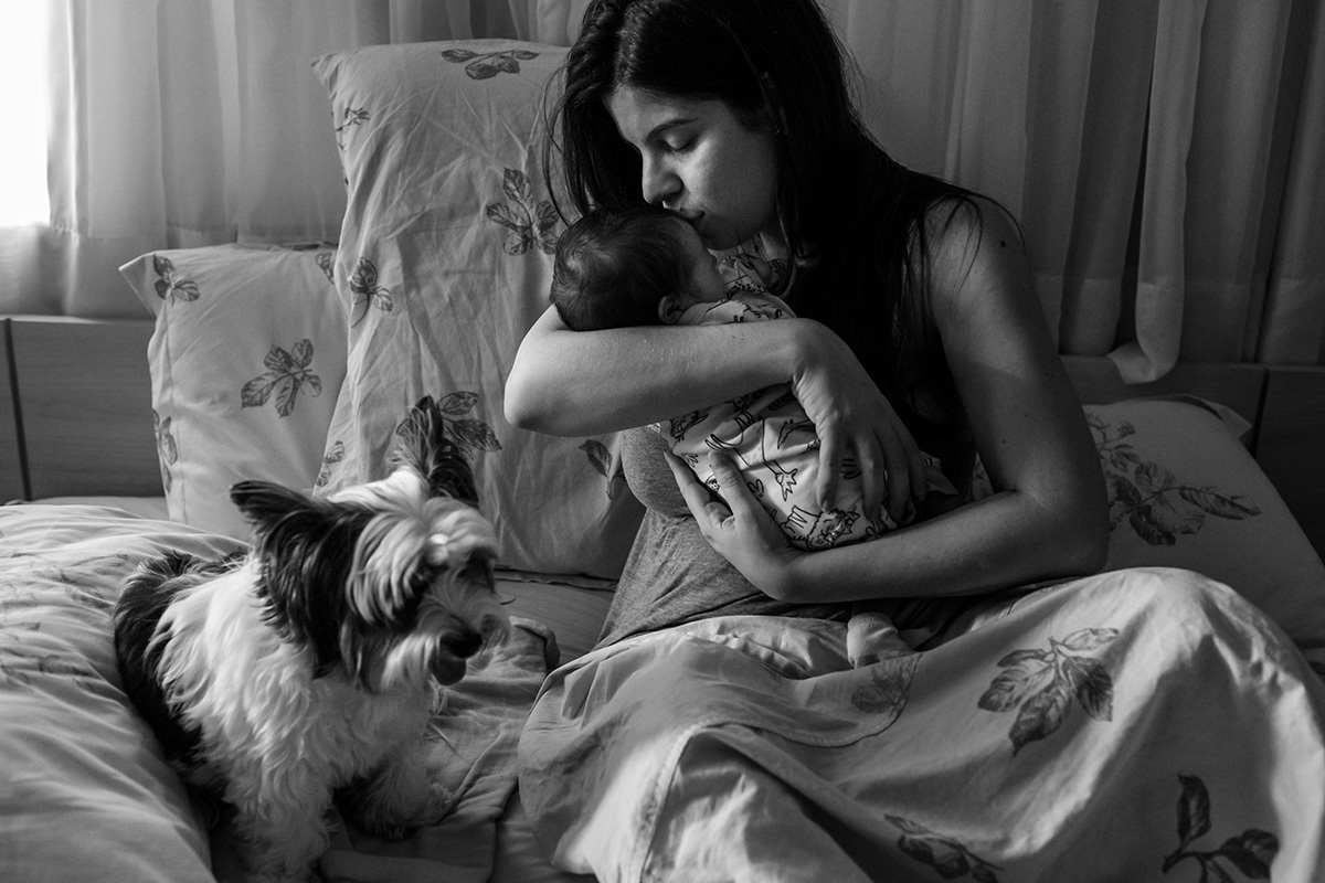 mãe beija seu bebê recém-nascido ainda na cama de casal ao acordar pela manhã, num registro documental feito por Adriana Costa