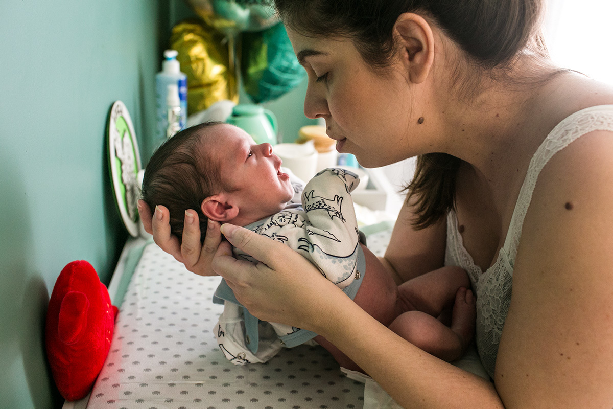 mãe olha delicadamente seu bebê recém-nascido nos olhos durante a troca de fralda, em foto de Adriana Costa