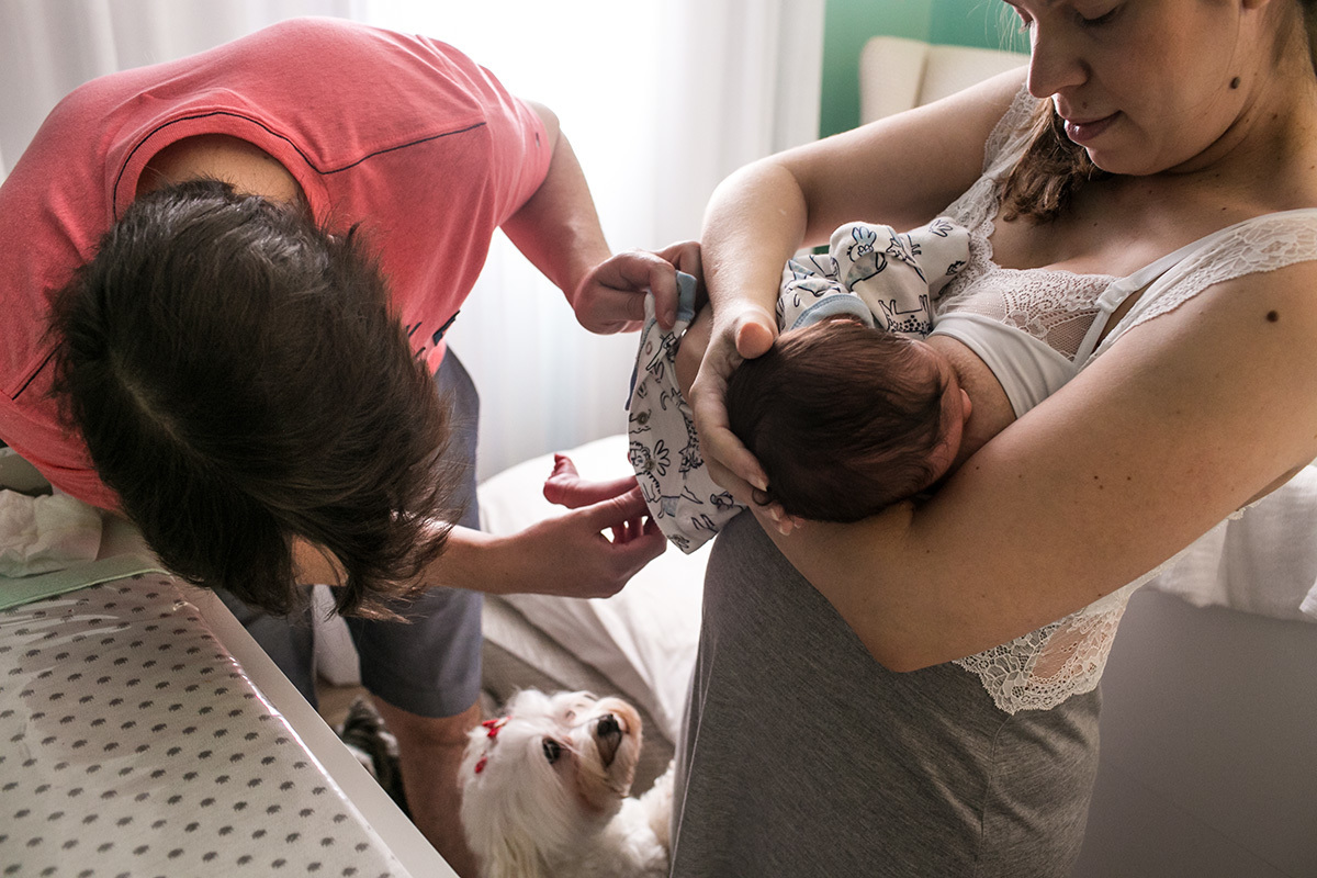 pai confere se o bumbum do seu bebê que está no colo da mãe está limpo e seu cãozinho observa tudo, por Adriana Costa