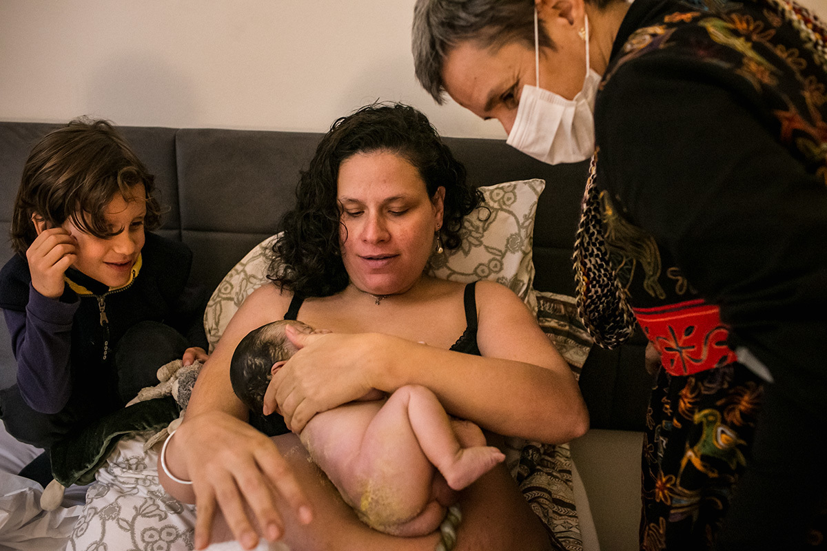 mãe ajeita seu bebê que acabou de nascer, ainda com vérnix em seu colo ao lado de seu filho mais velho e de sua enfermeira obstetra