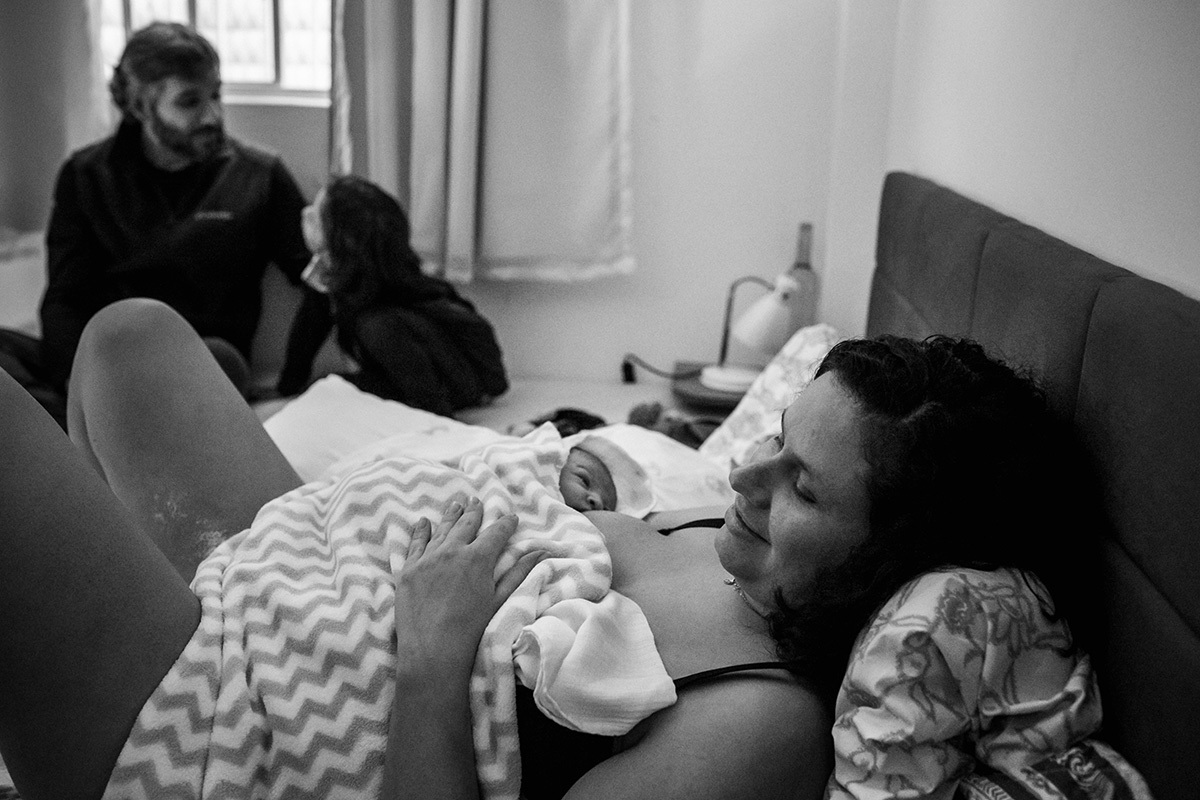 mãe descansa em sua cama com seu bebê após o parto ao lado de seu marido e filho mais velho, numa foto de Adriana Costa