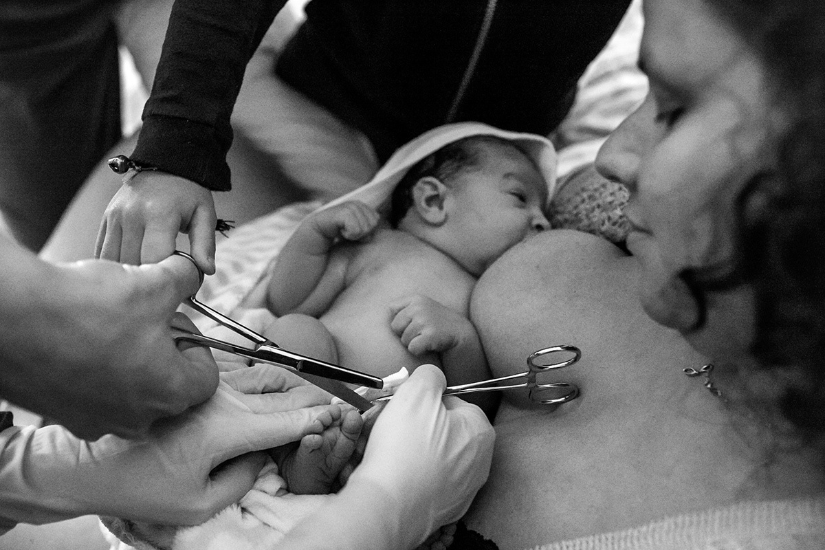 bebê tendo seu cordão umbilical cortado pelo pai e pelo irmão enquanto está amamentando, em foto de Adriana Costa