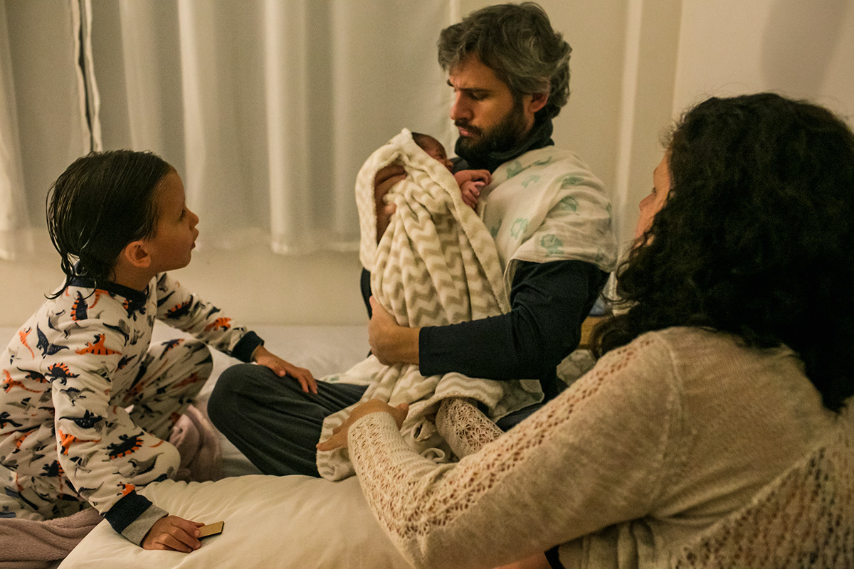 pai pega seu bebê que acabou de nascer nos braços ao lado de seu outro filho, em foto de Adriana Costa