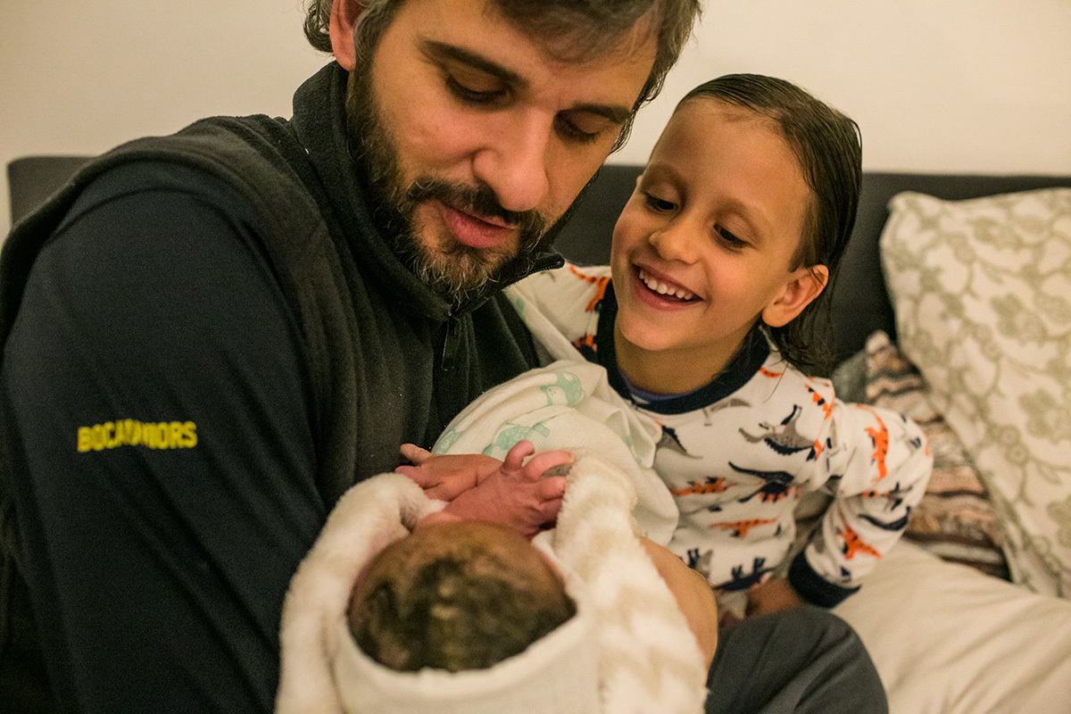 Foto de Adriana Costa de um pai admirando seu filho que acabou de nascer em seu colo ao lado de seu filho mais velho