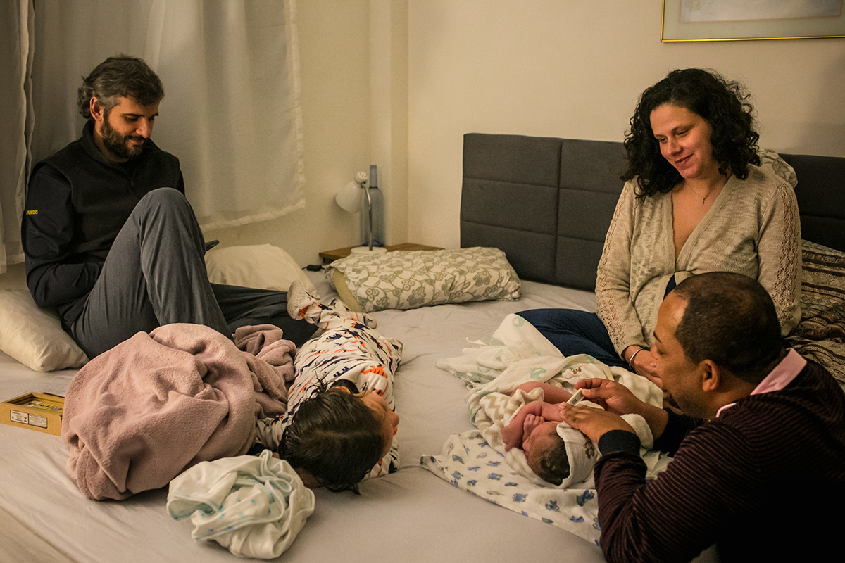 família reunida na cama do casal enquanto o enfermeiro obstetra avalia o bebê que acabou de nascer por um parto domiciliar, em foto de Adriana Costa