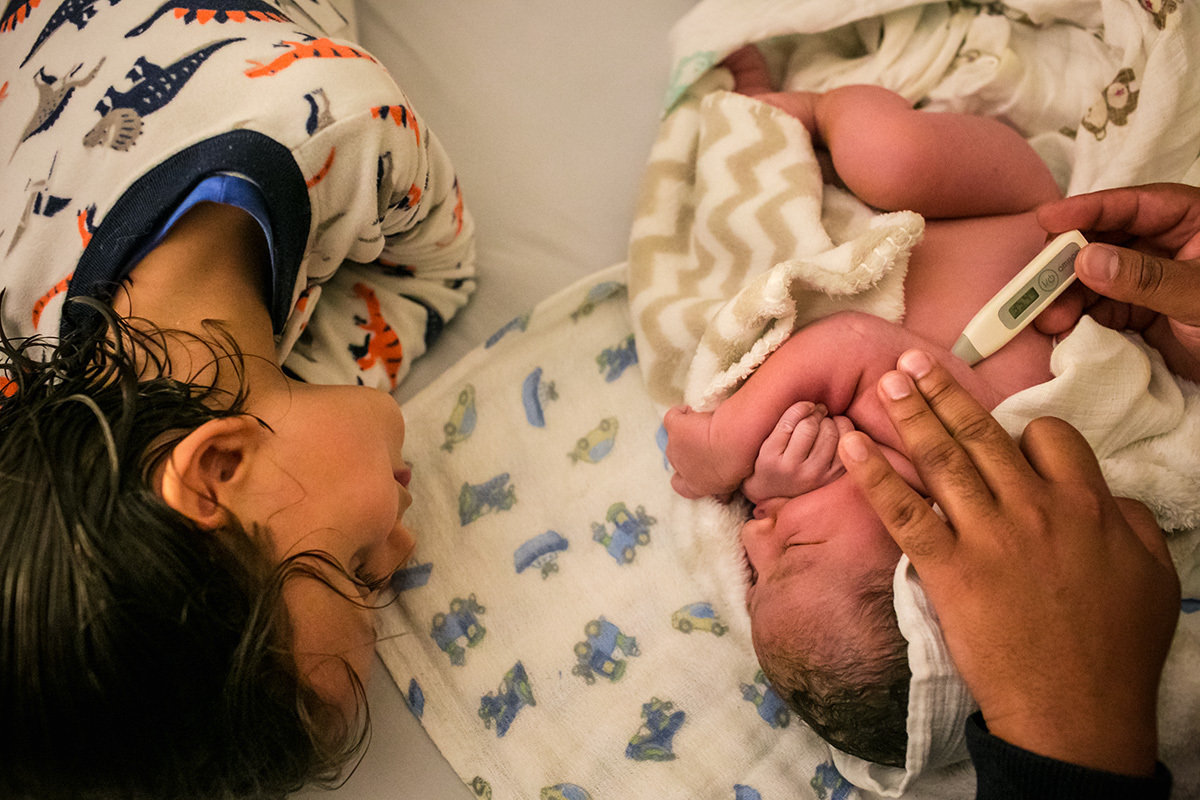 Adriana costa documentou menino observando  seu irmão que acabou de nascer ser examinado na cama de seus pais