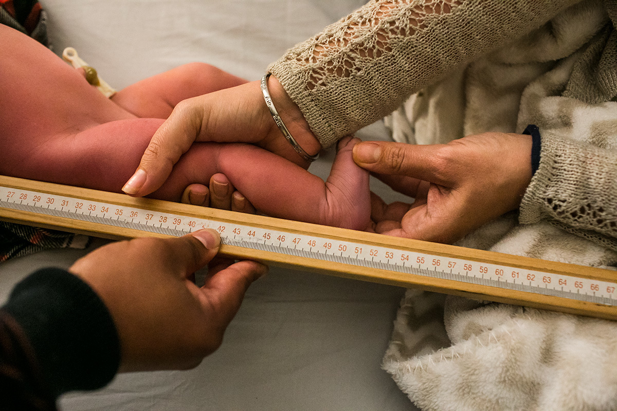 mãe ajudando a medir seu filho em seu primeiro exame após o nascimento, numa foto de Adriana Costa