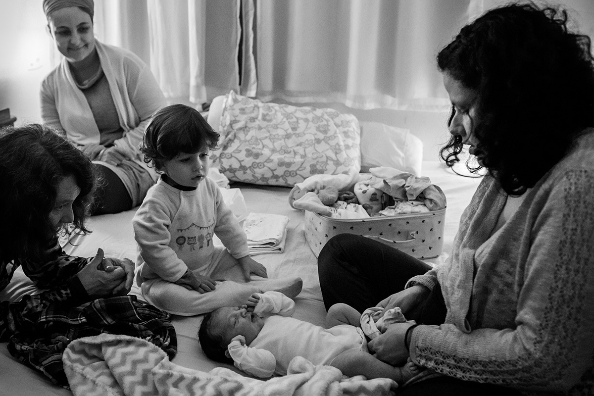 mãe colocando a roupa em seu bebê após o nascimento ao lado de seu outro filho, da doula e de sua mãe