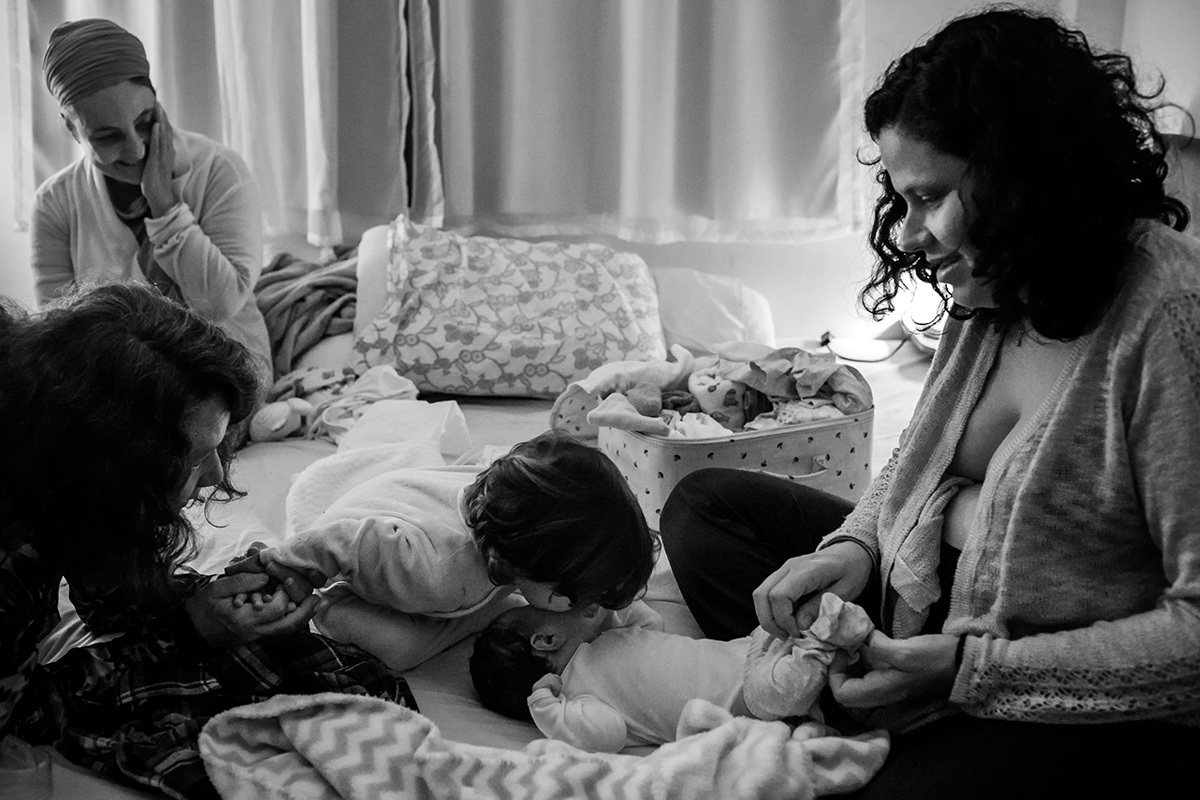 avó e doula riem do menino beijando seu irmão enquanto sua mãe o veste pela primeira vez após o nascimento, por Adriana Costa