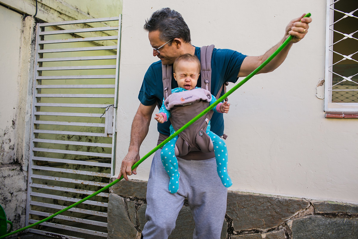 pai  com sua filha de um ano no canguru para bebês ajeitando a mangueira para molhar o jardim, por Adriana Costa