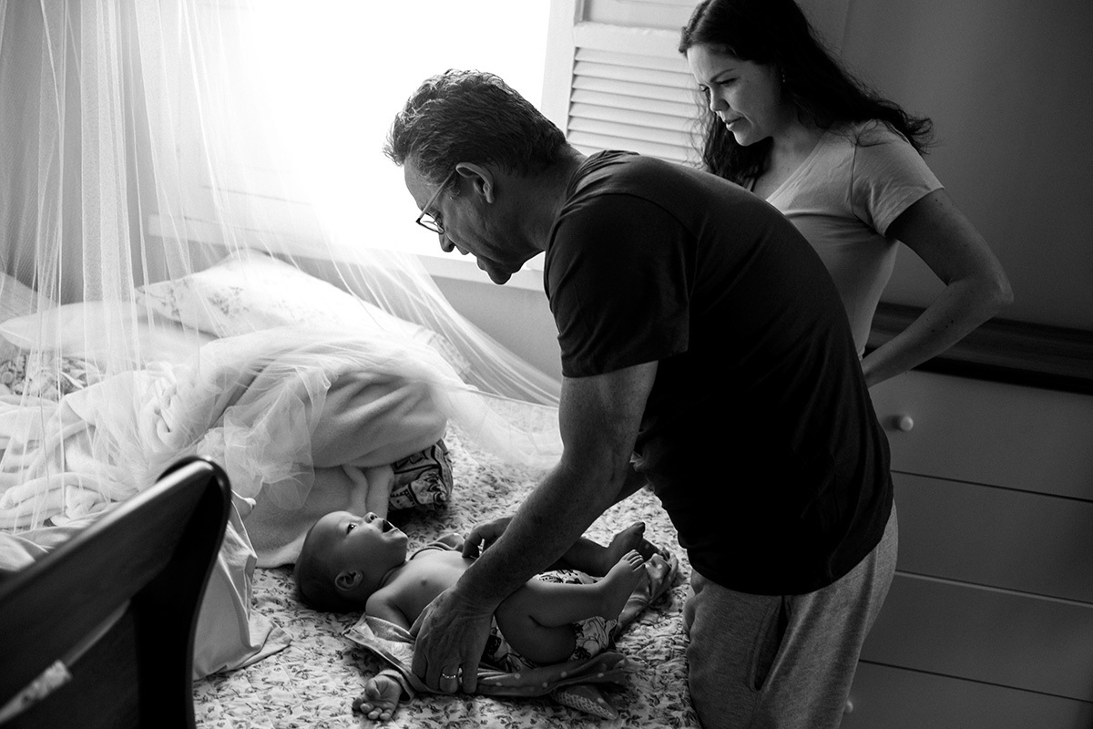 pais trocando a roupa da filha no quarto do casal, em foto de Adriana Costa