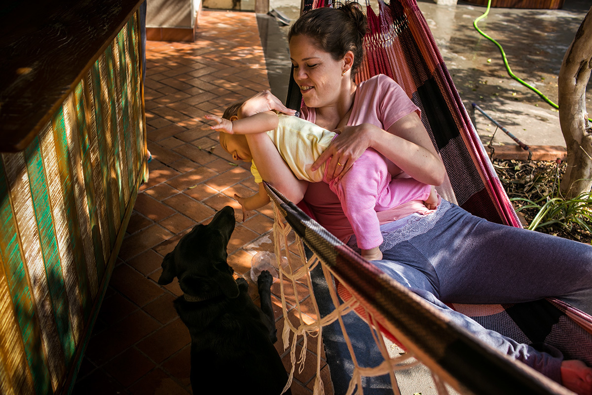Foto de Adriana Costa da mãe e sua filha de um ano deitadas na rede e brincando com o cachorro