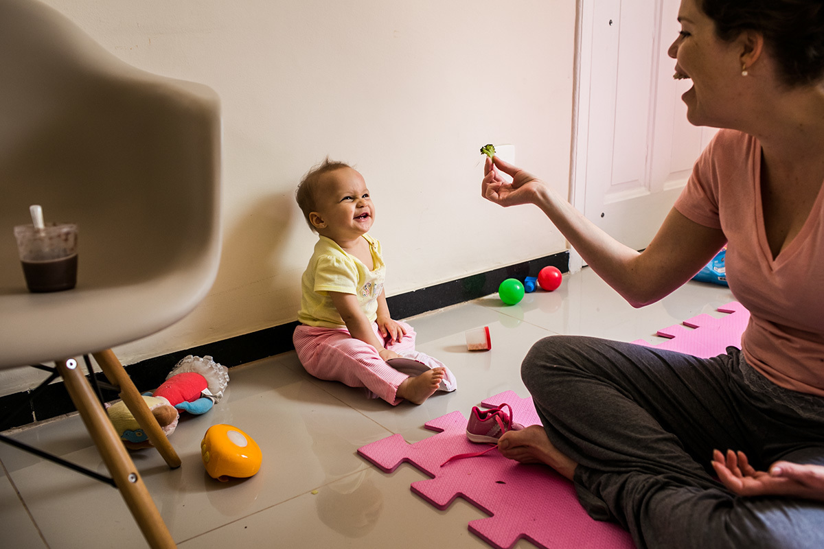 bebê olha super feliz para o brócolis na mão da mãe, em foto de Adriana Costa