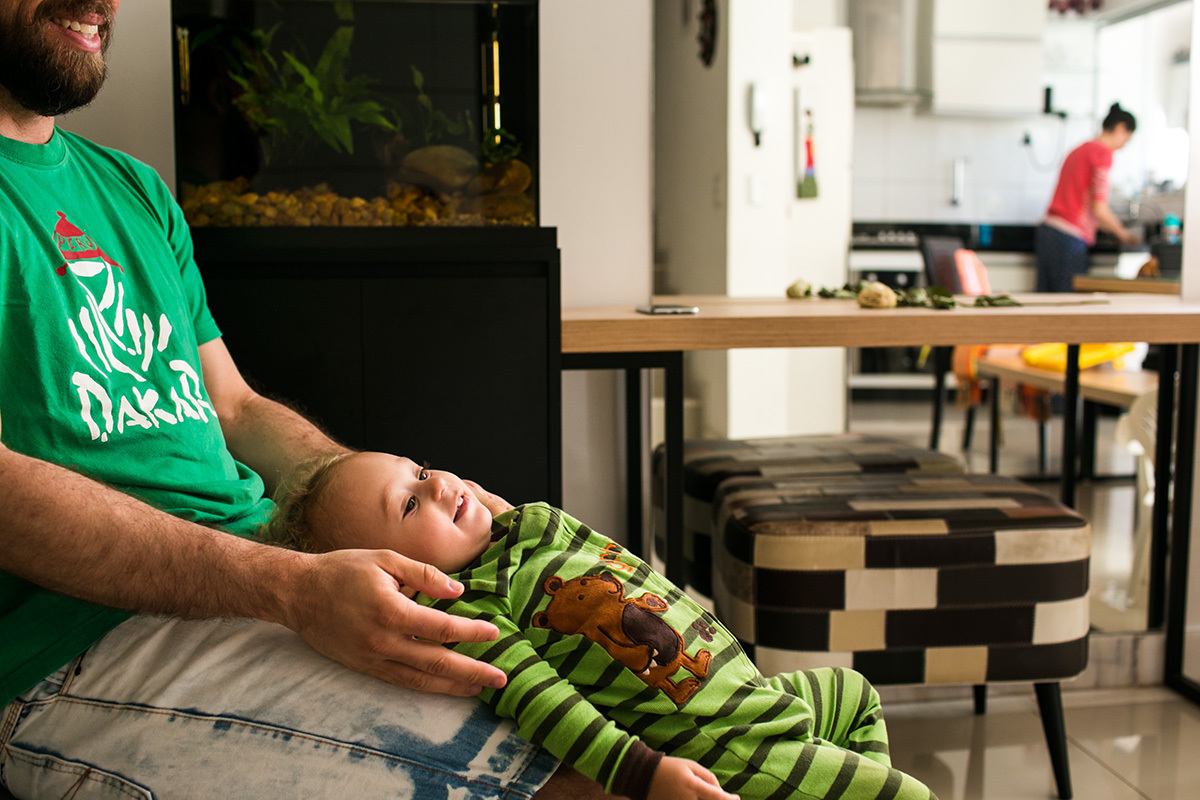 pai brinca de escorregar seu menino por suas pernas enquanto a mãe arruma a cozinha, em foto de Adriana Costa