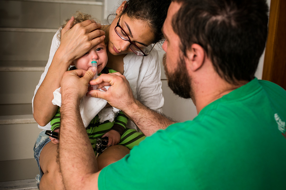 pai com a ajuda da mãe lava o nariz de seu bebê de um ano com soro fisiológico, em foto de Adriana Costa