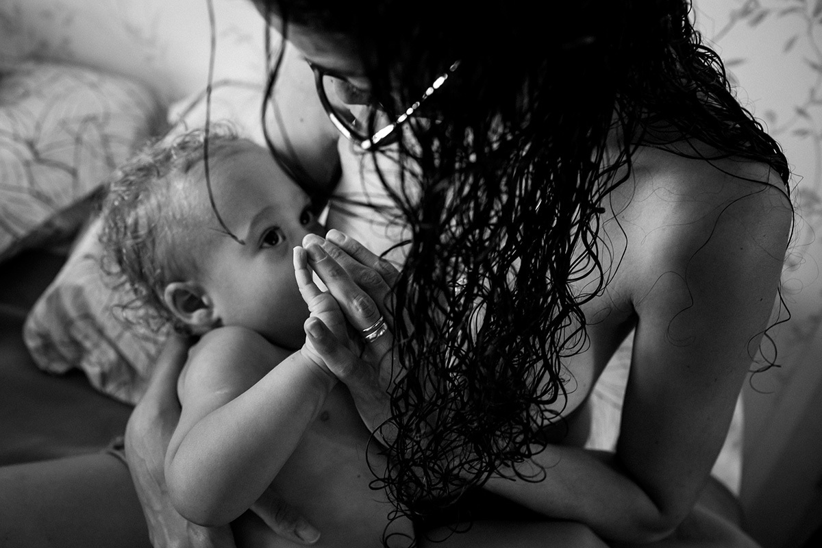 mãe amamenta seu filho de mãos dada com ele, em foto de Adriana Costa