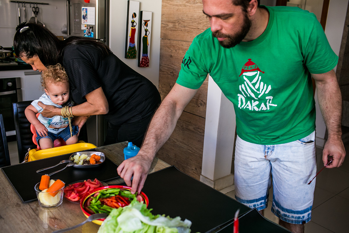 avó coloca o neto na cadeirinha para almoçar enquanto o pai pega um legume da mesa