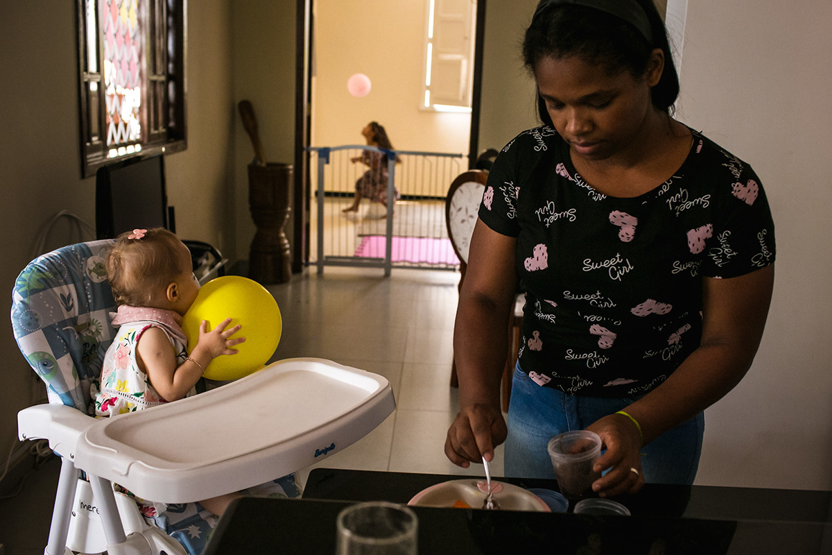 aniversariante vendo uma outra criança brincar com balão enquanto ela almoça segurando seu próprio balão, em foto de Adriana Costa