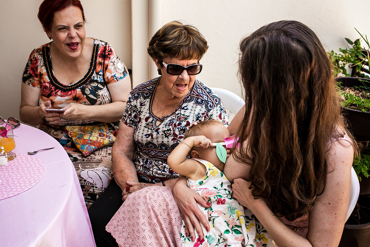 aniversariante de 1 ano amamentando e brincando de esconder seus olhos com a lembrancinha da festa, por Adriana Costa